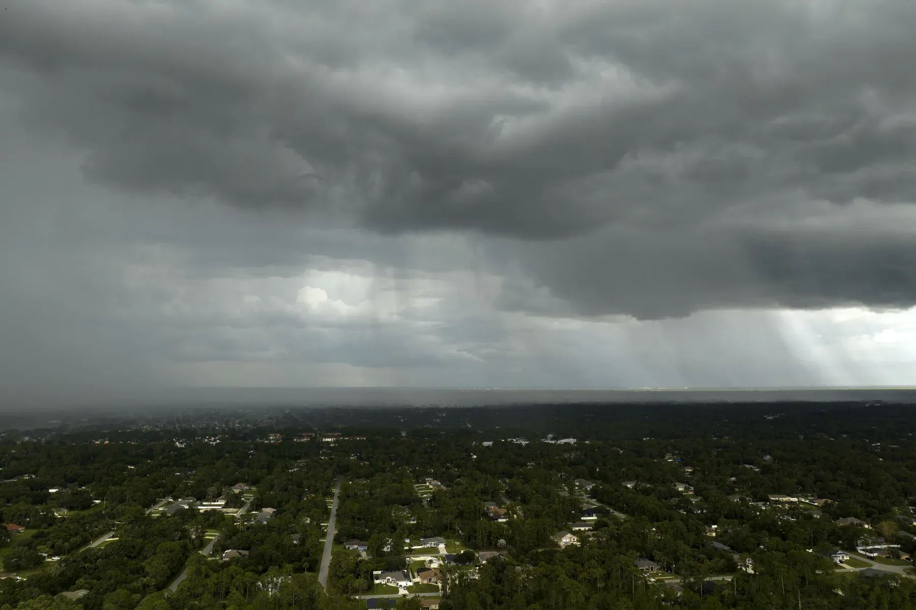 Dark storm clouds and rain shafts moving over a flat, tree-covered suburban neighborhood